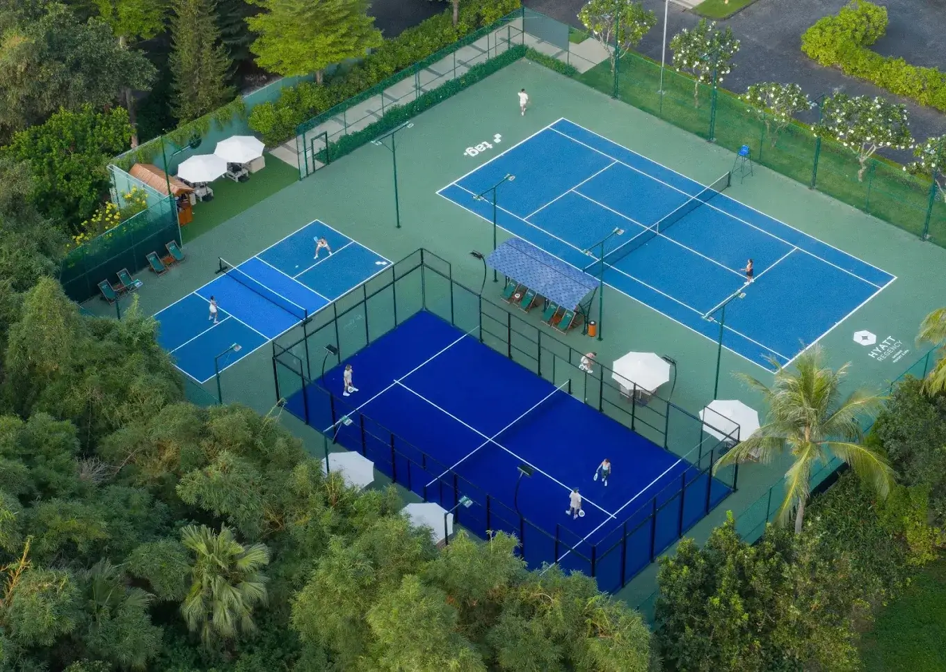Aerial view of the open-air blue padel court between a tennis court and two pickleball courts, with loungers and parasols alongside
