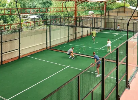 Two glass-walled padel courts side by side under a steel roof frame, four players mid-rally on the near court