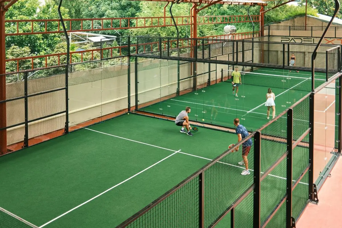 Two glass-walled padel courts side by side under a steel roof frame, four players mid-rally on the near court