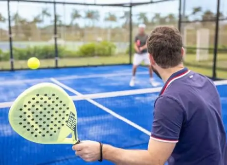 A player at the net of an open-air blue padel court, racket raised, with an opponent across the net and palm trees behind