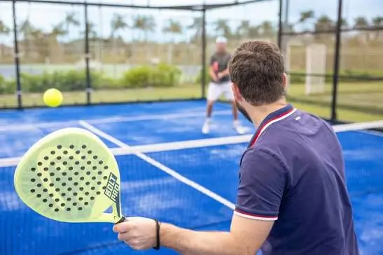 A player at the net of an open-air blue padel court, racket raised, with an opponent across the net and palm trees behind
